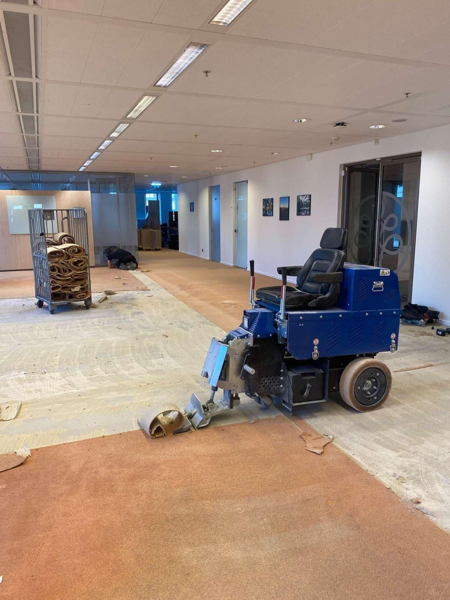 Kantoor waar het oude tapijt met een machine wordt verwijderd en een persoon op de vloer werkt.